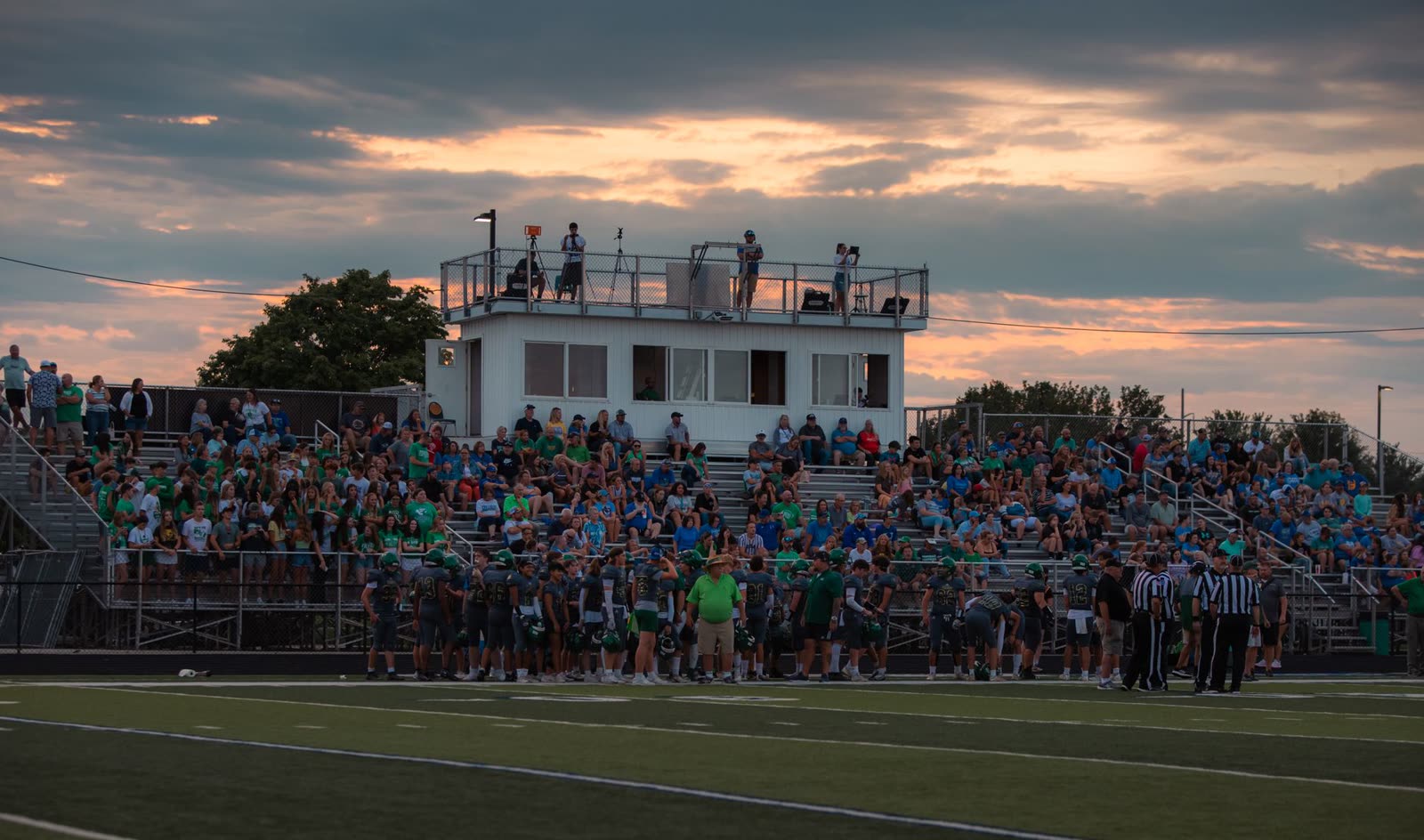 fans in the stands at a high school football game on a mild night / high school football stadium lights glowing at dusk in autumn / spectators in light sweatshirts watching a night football game