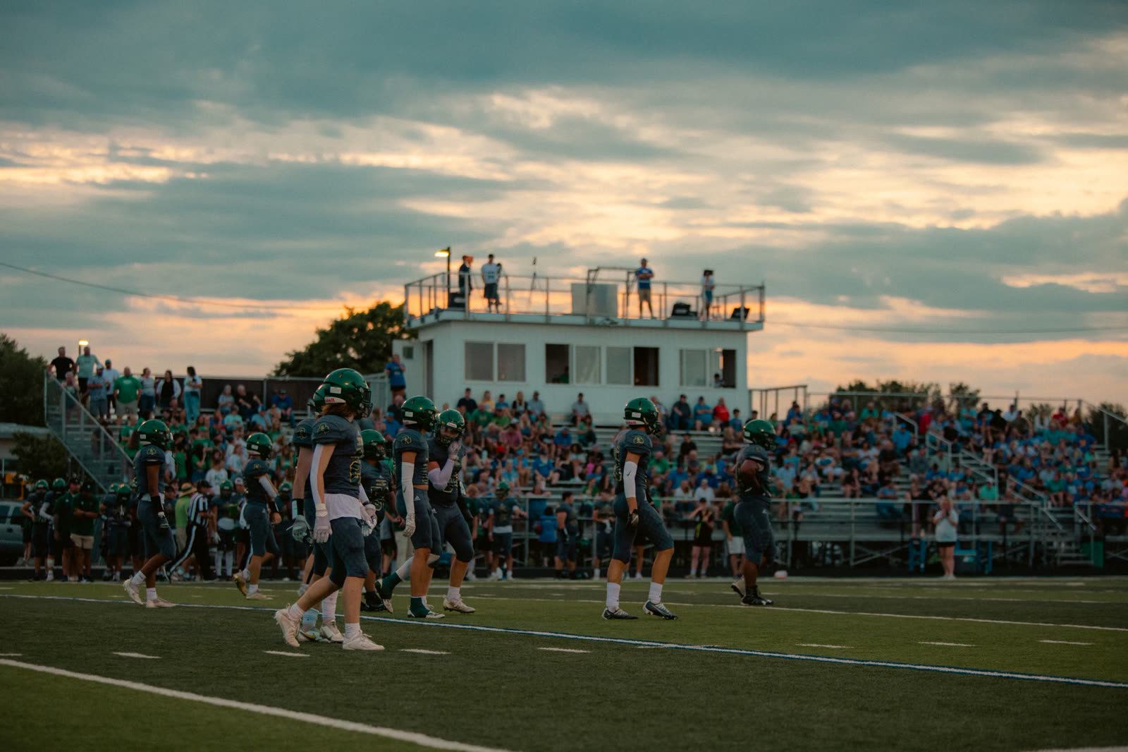 fans in short sleeves in high school football bleachers at night / high school football stadium lights glowing on a mild evening / crowd watching a night game from metal bleachers in autumn